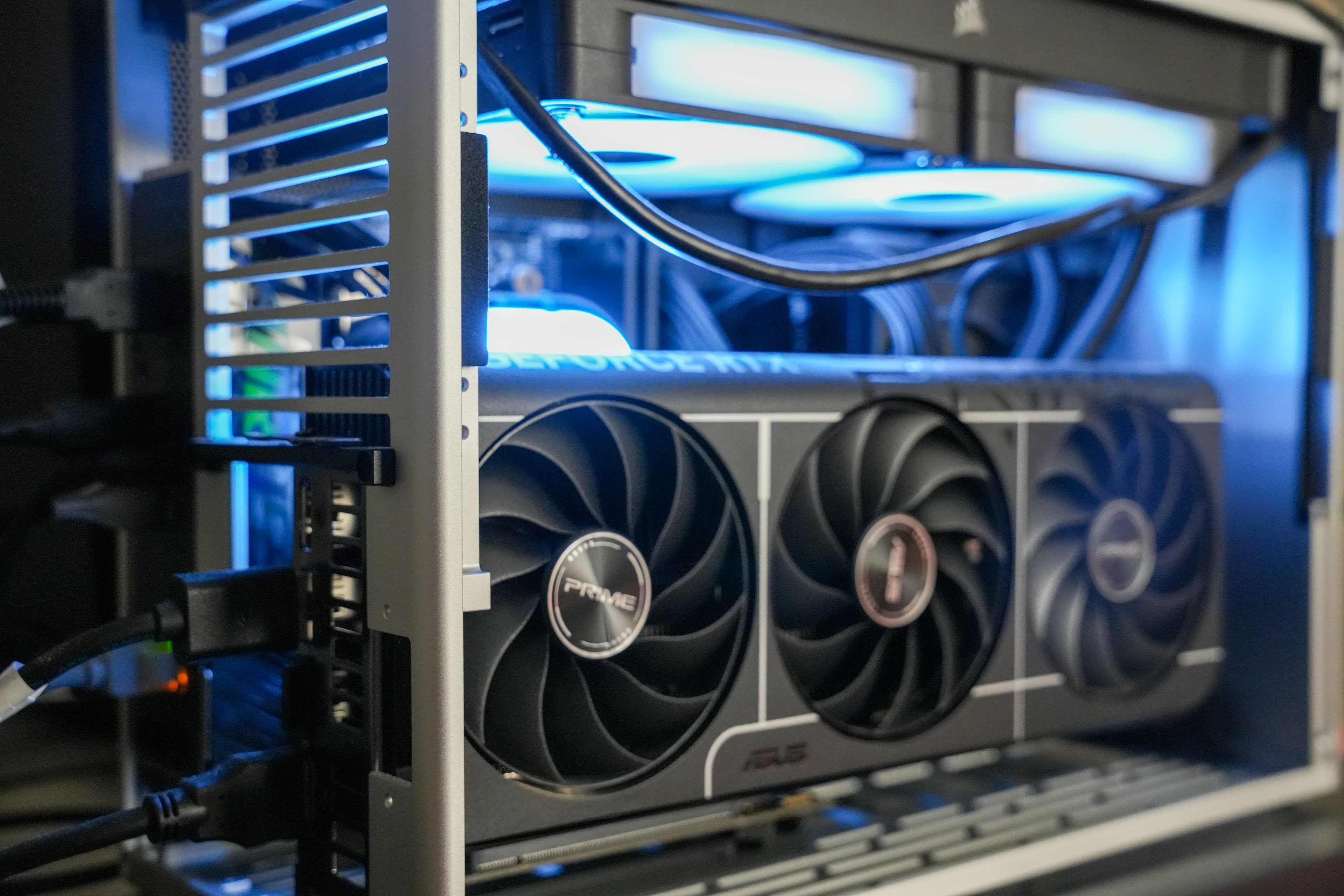 Close view of a triple-fan graphics card mounted horizontally in an open aluminium frame, with liquid-cooling tubing and a blue-lit radiator above it.